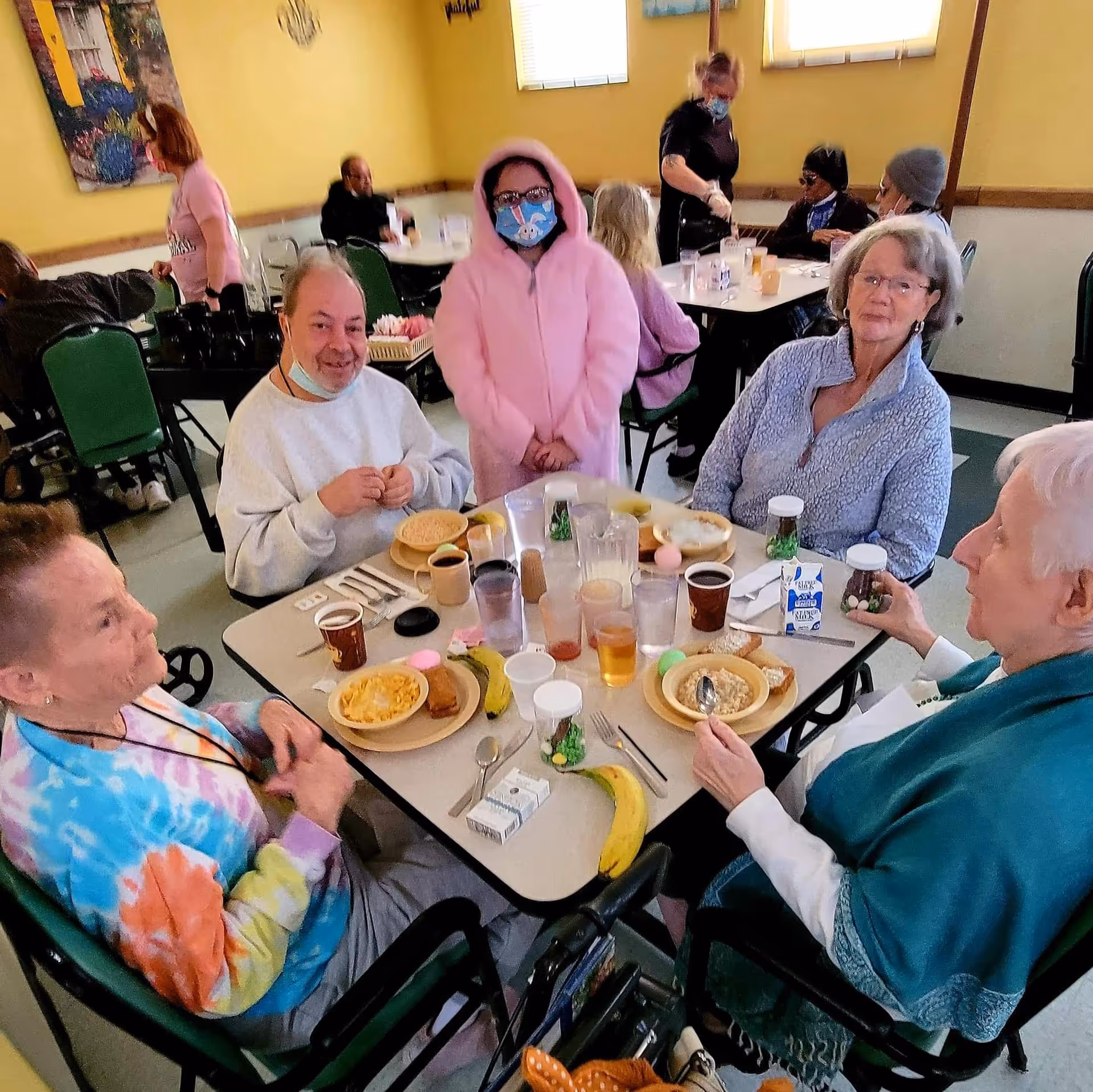 A group of senior residents and a staff member wearing masks sit around a table eating breakfast in a communal dining room.