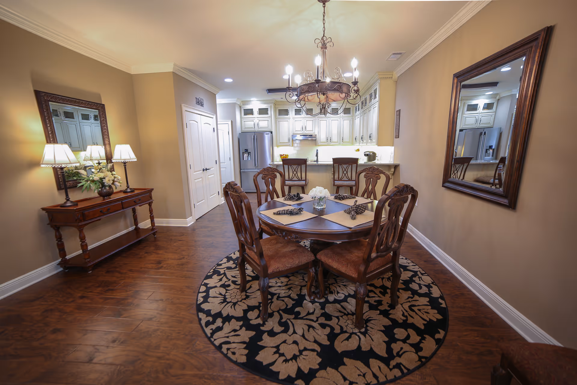 A dining area with a round wooden table set with placemats and napkins, surrounded by four wooden chairs with cushioned seats. Above the table hangs an ornate chandelier. The room has wooden flooring with a large floral patterned round rug under the table. To the left is a wooden console table with two lamps and a flower arrangement, and a large mirror above it. In the background is a kitchen area with white cabinets, a refrigerator, and a countertop with two bar stools. Another large mirror is mounted on the right wall.