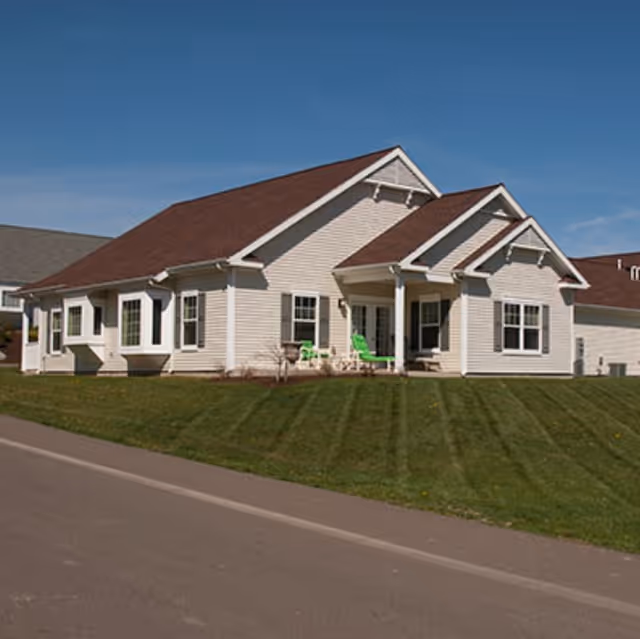A single-story residential building with beige siding and a brown roof under a clear blue sky. The building has multiple windows and a small covered porch with green chairs on a well-maintained lawn.