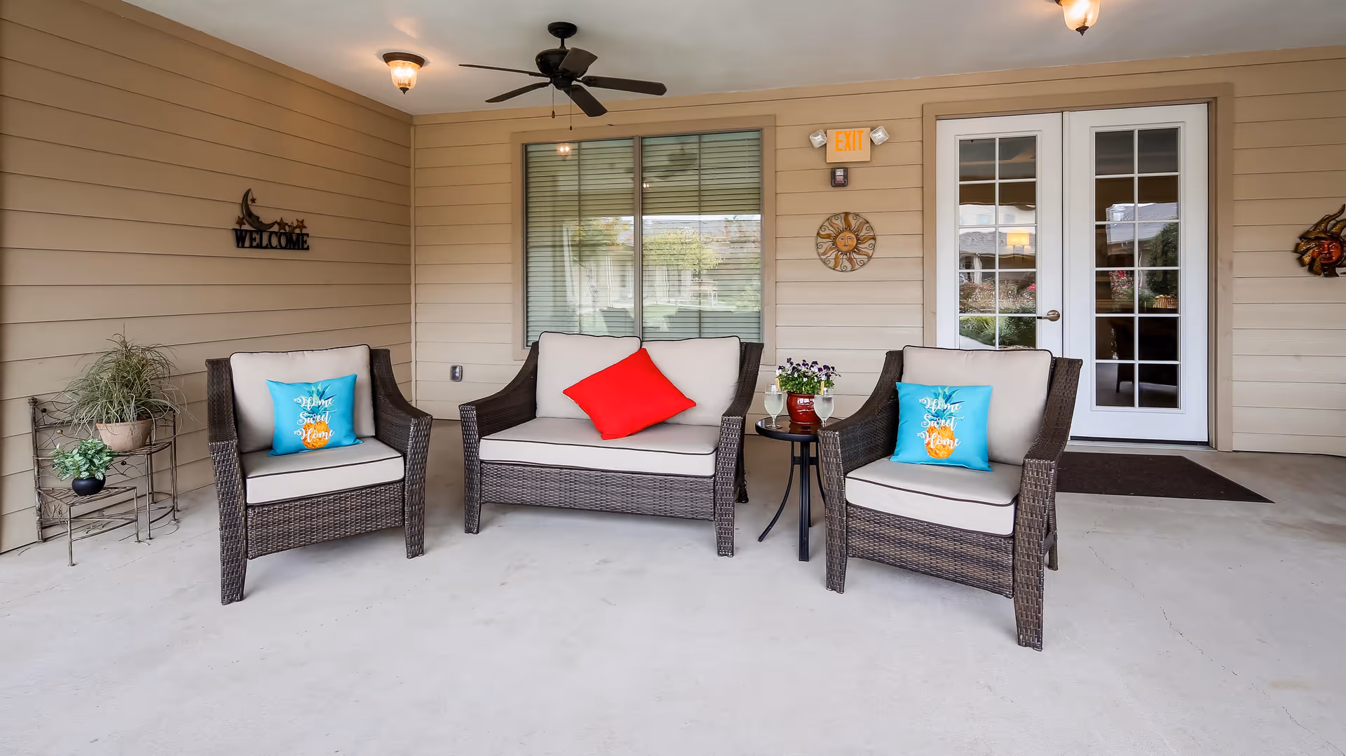 Outdoor covered patio area with three wicker chairs and a loveseat, all with beige cushions. Two chairs have blue pillows with orange designs and the loveseat has a red pillow. A small round table between the loveseat and one chair holds a potted plant and two glasses of white wine. The wall behind features a window with blinds, a door with glass panes, a ceiling fan, a 'Welcome' sign, and decorative wall hangings.