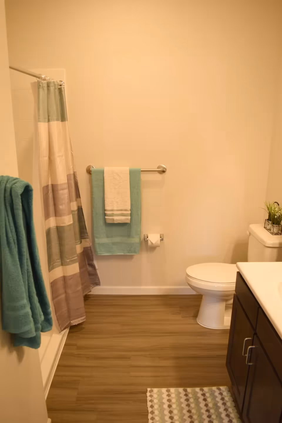 A bathroom with a white toilet, a dark wood vanity with a white countertop, a shower with a striped shower curtain in shades of green, beige, and brown, and two towel racks holding teal and white towels. The floor is wood-style laminate, and there is a small patterned rug in front of the vanity.