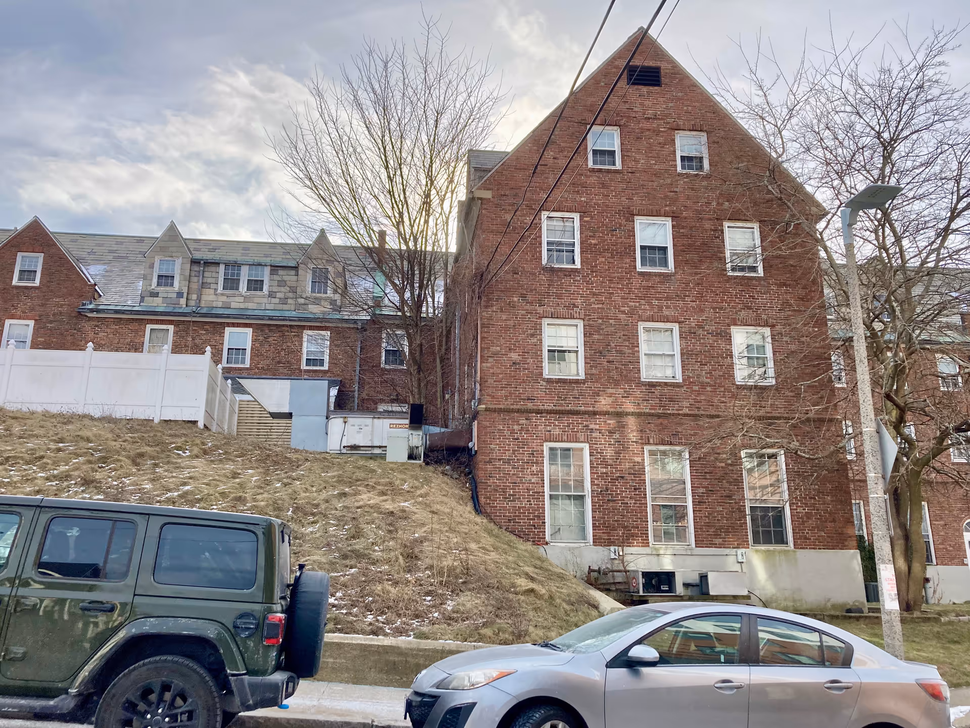 Exterior view of a multi-story red brick building with multiple windows, a sloped grassy area in front, two parked cars, and leafless trees under a cloudy sky.