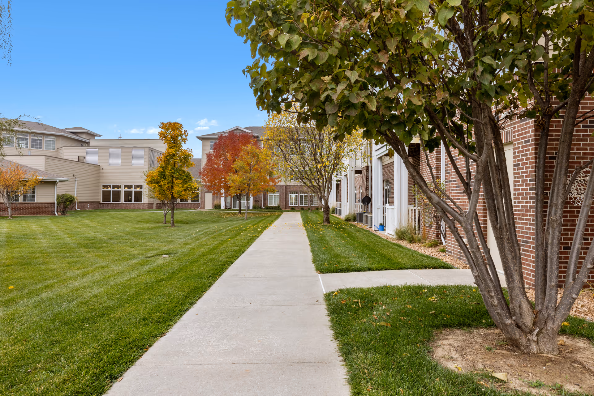 A paved walkway runs through a well-maintained grassy courtyard with trees showing autumn colors, surrounded by a senior living facility building with brick and beige siding under a clear blue sky.