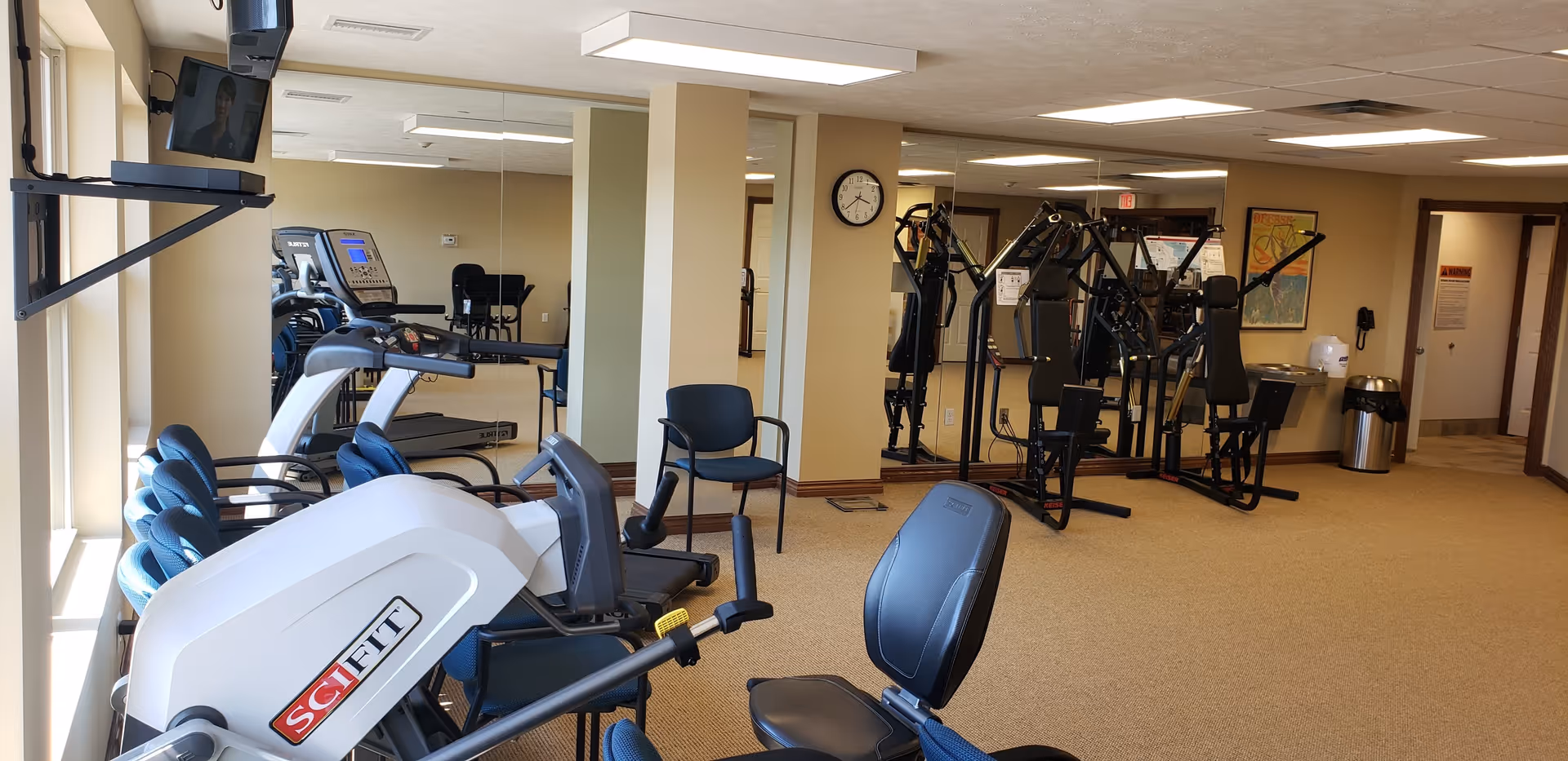 Interior view of a fitness room in a senior living facility with exercise equipment including a SciFit machine, treadmills, and weight machines. There are several blue chairs along the window, a wall clock, a mounted TV, and a water fountain in the background. The walls are beige and there are large mirrors on one side.
