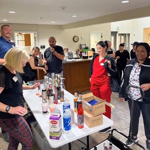 A group of people gathered around a table with various drink syrups, bottles, and mixers in a room with a reception desk in the background. Some individuals are smiling and interacting, and one person is using a walker.