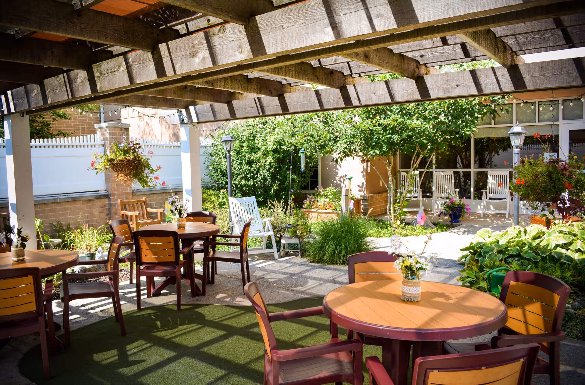 Shaded outdoor courtyard with round tables and chairs under a wooden pergola surrounded by plants and greenery.