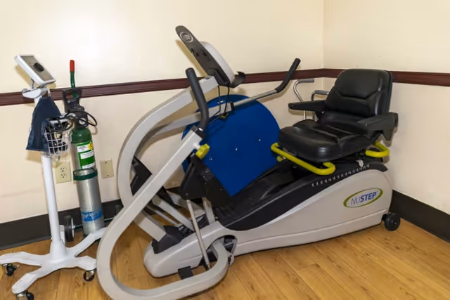 A NuStep exercise machine with a black cushioned seat and handles, placed on a wooden floor against a beige wall with a brown trim. Next to the machine is a medical oxygen tank on a wheeled stand with a digital monitor attached.