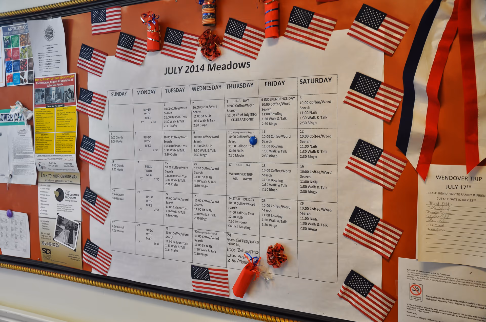 A bulletin board decorated with small American flags and a large calendar titled 'JULY 2014 Meadows' showing daily activities and events. The calendar includes activities such as coffee/word search, balloon toss, walk & talk, bingo, and special events like Independence Day and a Wendover trip. Additional notices and flyers are pinned to the left and right sides of the board.