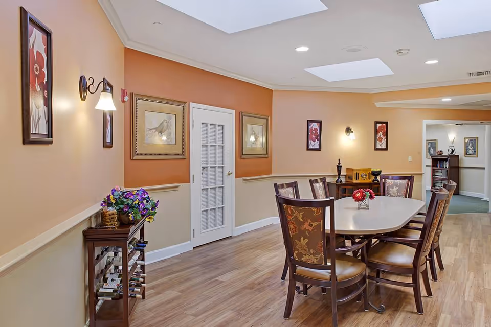 A dining room with a rectangular table surrounded by six chairs with floral upholstery. The walls are painted in warm tones with framed artwork and wall sconces. There is a small wooden wine rack with bottles and a floral arrangement on top. The room has a wooden floor and a ceiling with recessed lighting and skylights.
