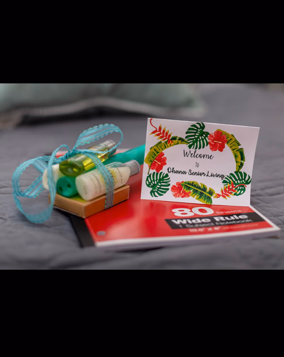 A welcome card with tropical leaf and flower designs that reads 'Welcome to Ohana Senior Living' placed on top of a red wide rule notebook with 80 sheets. Next to the card are small toiletry bottles tied together with a blue ribbon, all set on a gray quilted surface.