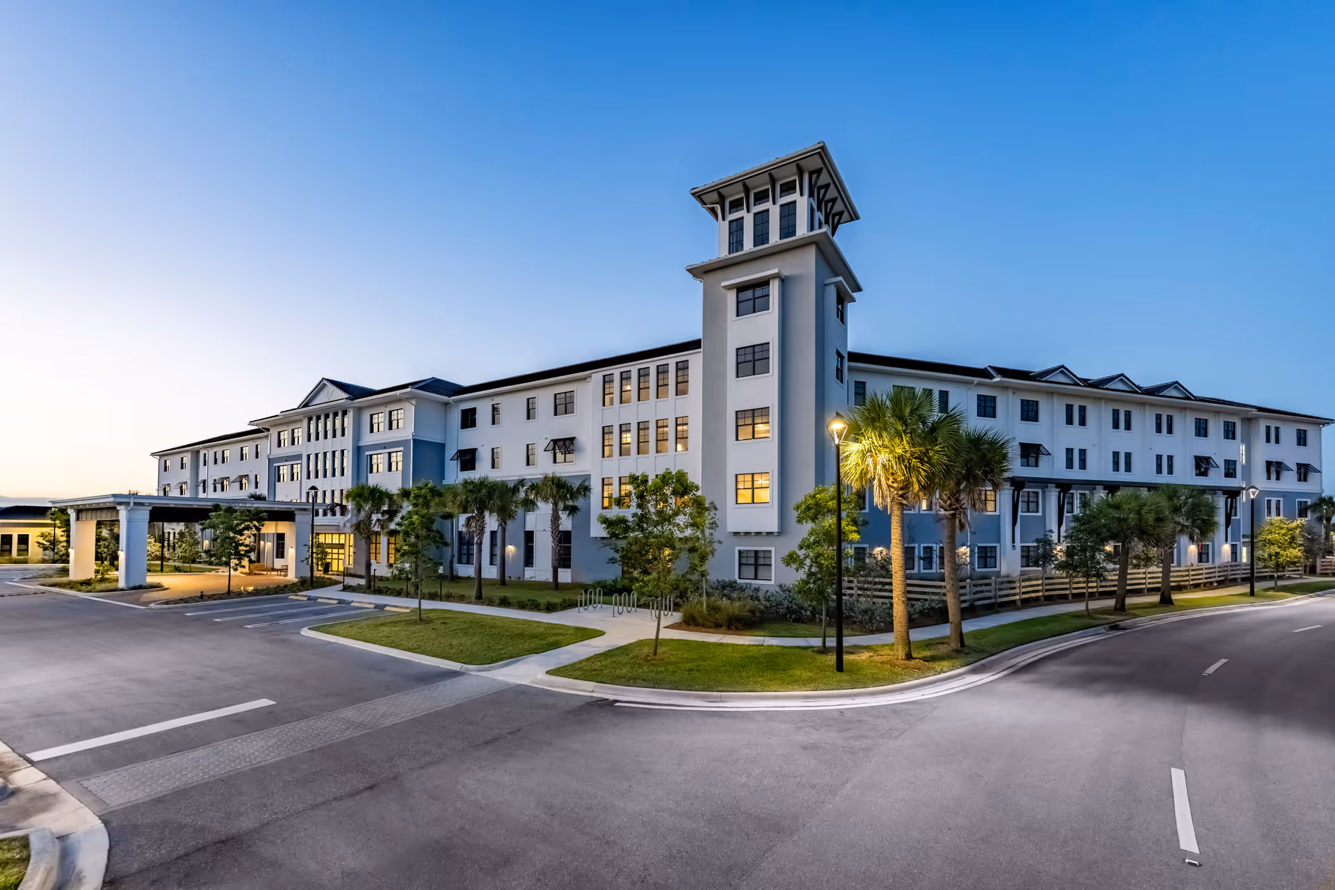 Exterior front view of a large multi-story senior living building at dusk with palm trees and a covered entrance.