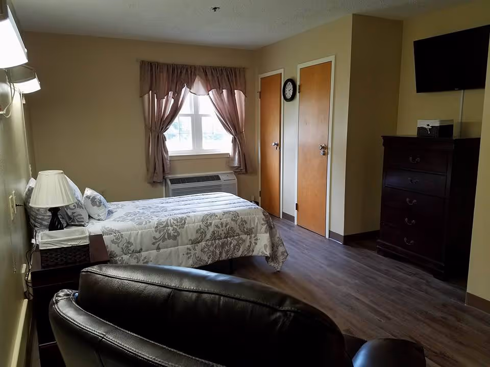 A senior living facility bedroom with a single bed covered in a patterned bedspread, a bedside table with a lamp and tissue box, a window with curtains, two closed wooden doors, a wall clock, a dark wooden dresser, a mounted flat-screen TV, and a leather chair in the foreground.