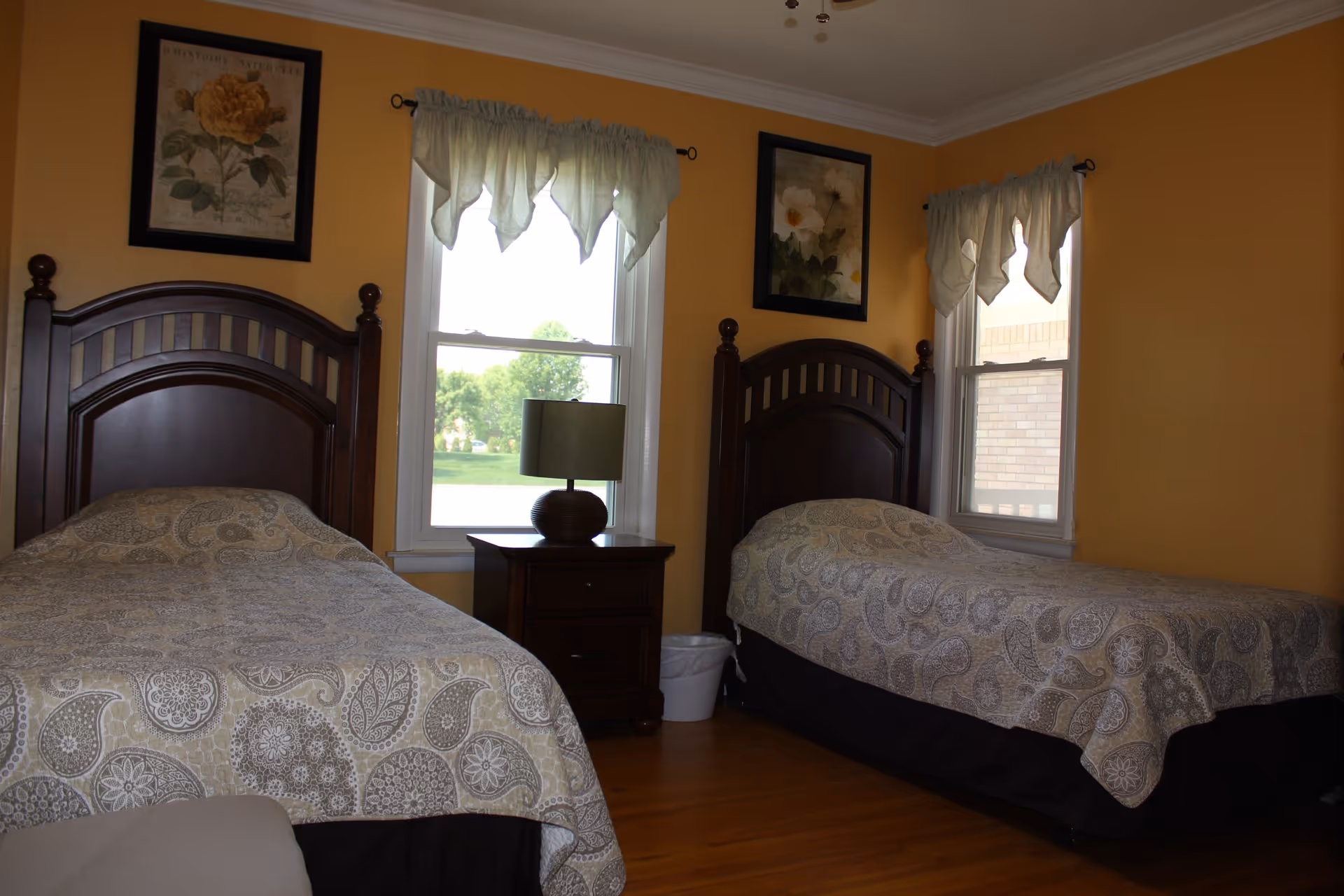 A bedroom with two twin beds featuring dark wooden headboards and beige paisley-patterned bedspreads. Between the beds is a dark wooden nightstand with a round lamp on top. Two windows with light-colored valances allow natural light into the room. The walls are painted yellow and decorated with framed floral artwork above each bed. The floor is wooden.