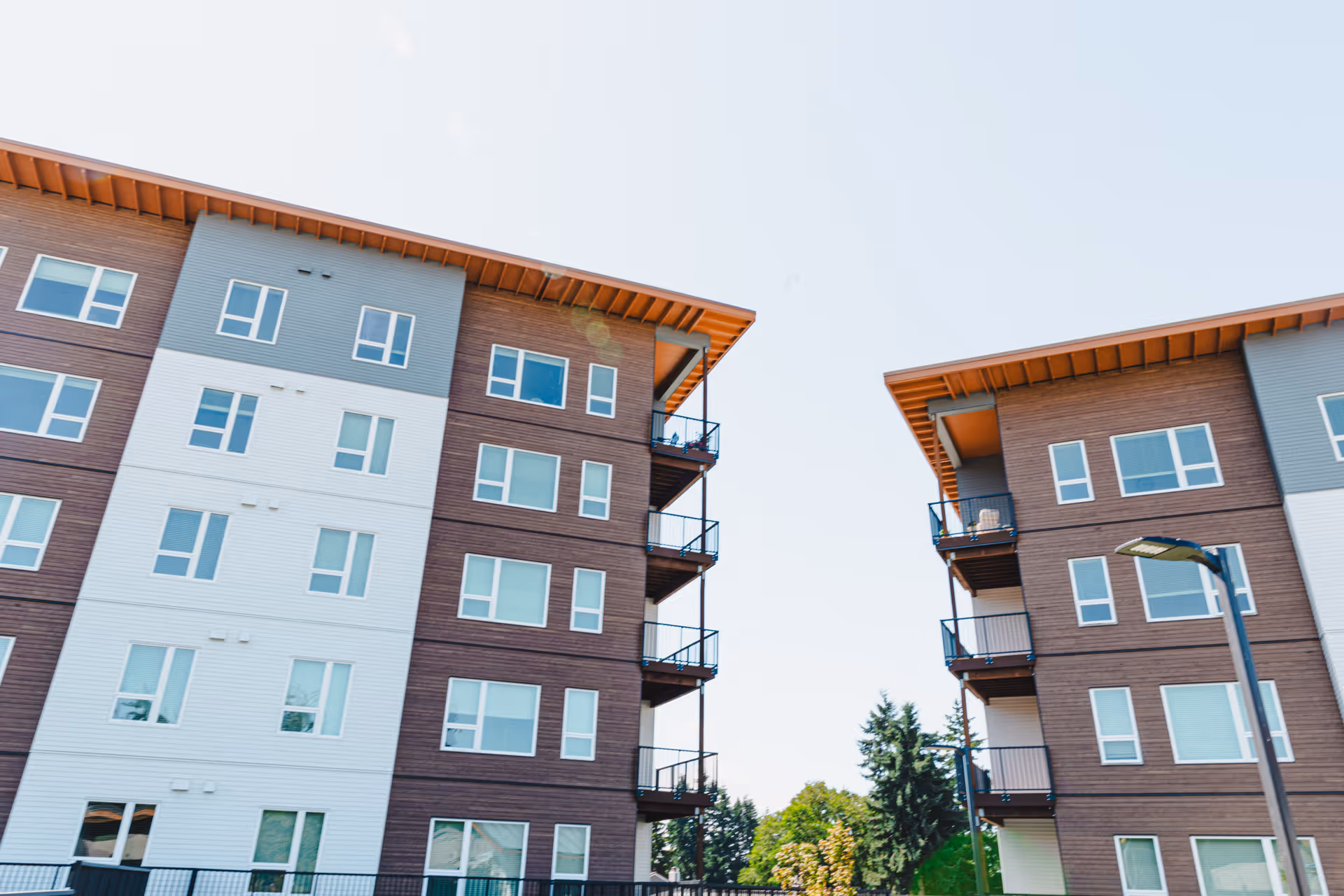 Exterior view of two modern multi-story residential buildings with balconies, set against a clear blue sky and some trees in the background.