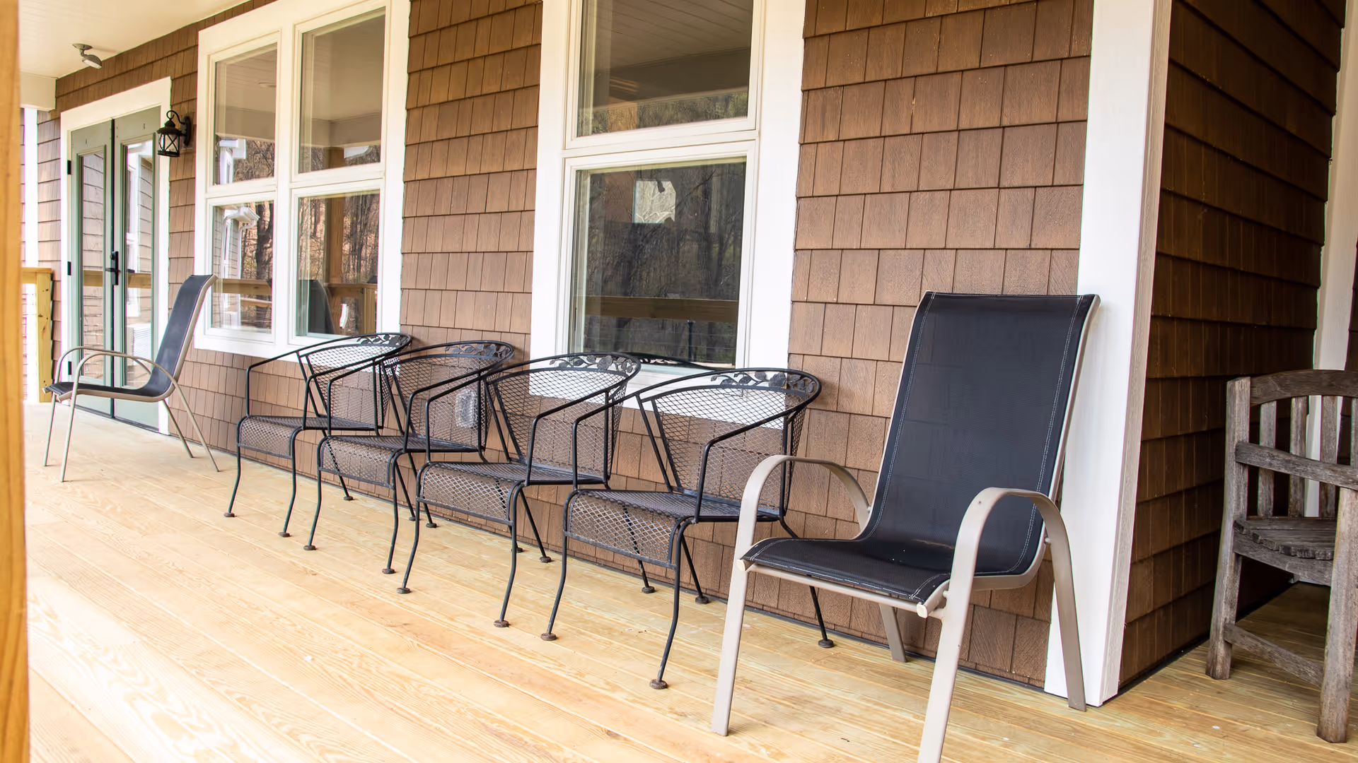 A wooden porch with several chairs lined up against the exterior wall of a building with brown shingle siding and white trim around the windows and door.