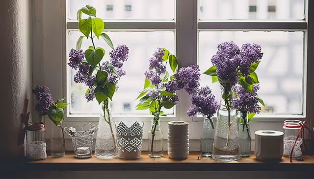 A windowsill lined with glass vases and jars holding sprigs of purple lilacs and decorative items.