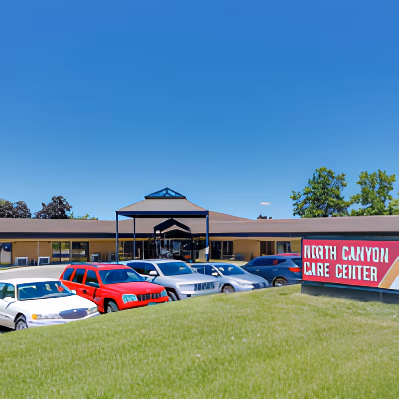 Exterior view of a single-story care center building with a covered entrance and several parked cars in front. There is a sign on the right side that reads 'North Canyon Care Center'. The sky is clear and blue, and there is green grass in the foreground.