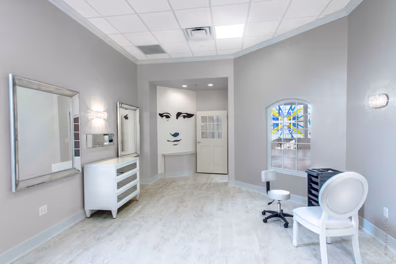 A bright, modern interior room with light gray walls and light wood flooring. The room features two large mirrors on the left wall above a white dresser, a white swivel chair and a small rolling stool on the right, and a window with a colorful stained glass design. At the far end, there is a white door and a wall with a minimalist black and white face artwork. The ceiling has recessed lighting and a grid pattern.