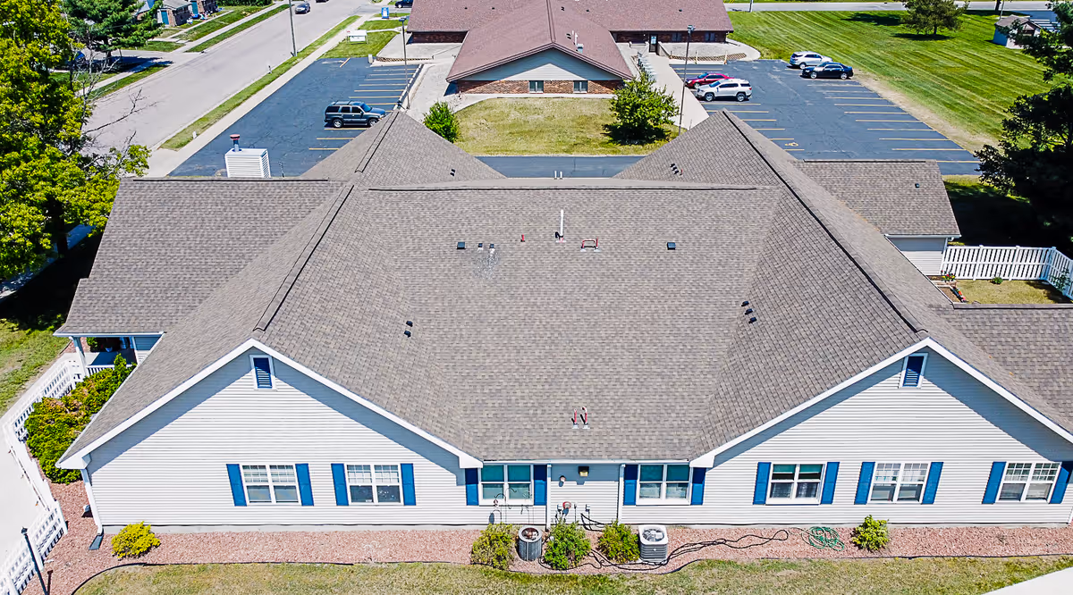 Aerial view of a single-story senior living facility building with a gray shingled roof and white siding with blue window shutters. The building is surrounded by a parking lot with several cars and green lawns. There are sidewalks and some small bushes around the building.