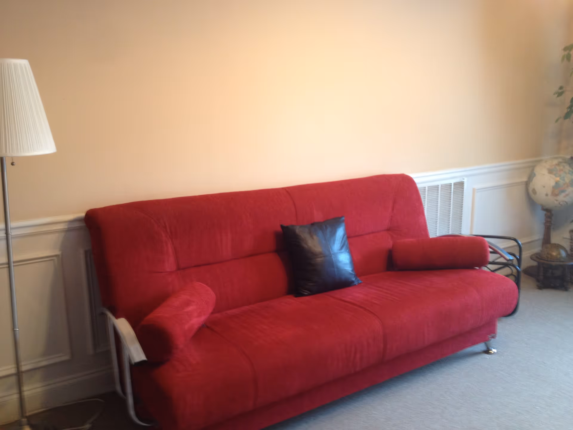 A red sofa with a black cushion and two cylindrical red pillows in a room with beige walls and white wainscoting. To the left of the sofa is a tall floor lamp with a white lampshade. To the right, there is a globe on a stand and a plant partially visible.