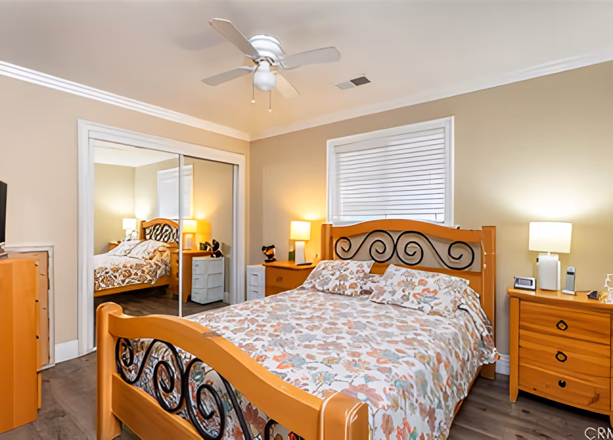 A cozy bedroom with a wooden bed frame featuring black decorative swirls, a floral patterned bedspread, two wooden nightstands with lamps, a window with white blinds, a ceiling fan, and a mirrored sliding closet door reflecting part of the room.