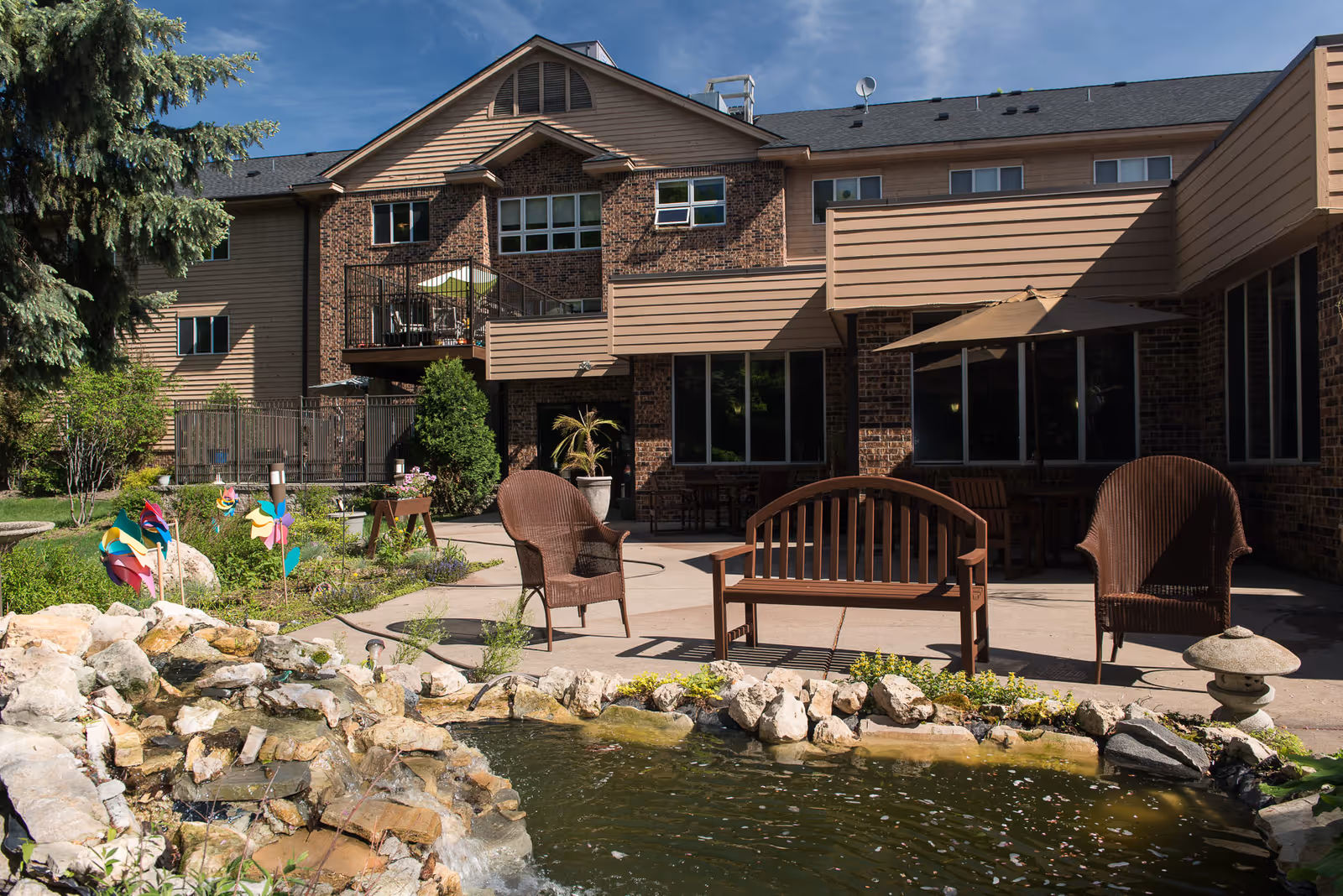 Outdoor courtyard with a small pond, benches, wicker chairs and umbrellas in front of a multi-story brick building.