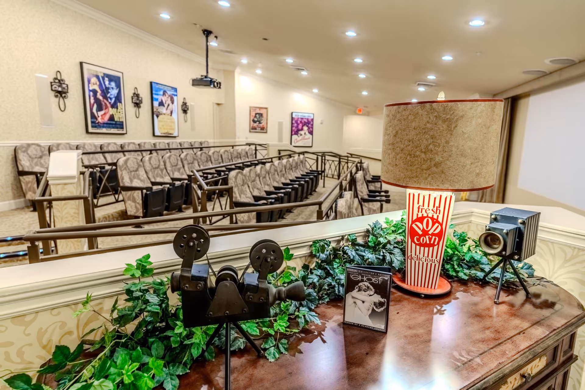 Interior view of a small theater room with rows of cushioned seats facing a large screen. The walls are decorated with vintage movie posters. In the foreground, there is a wooden table with decorative items including a vintage film projector, a popcorn-themed lamp, a small camera model, and a DVD case of the movie Ghost. Greenery decorates the table edge.