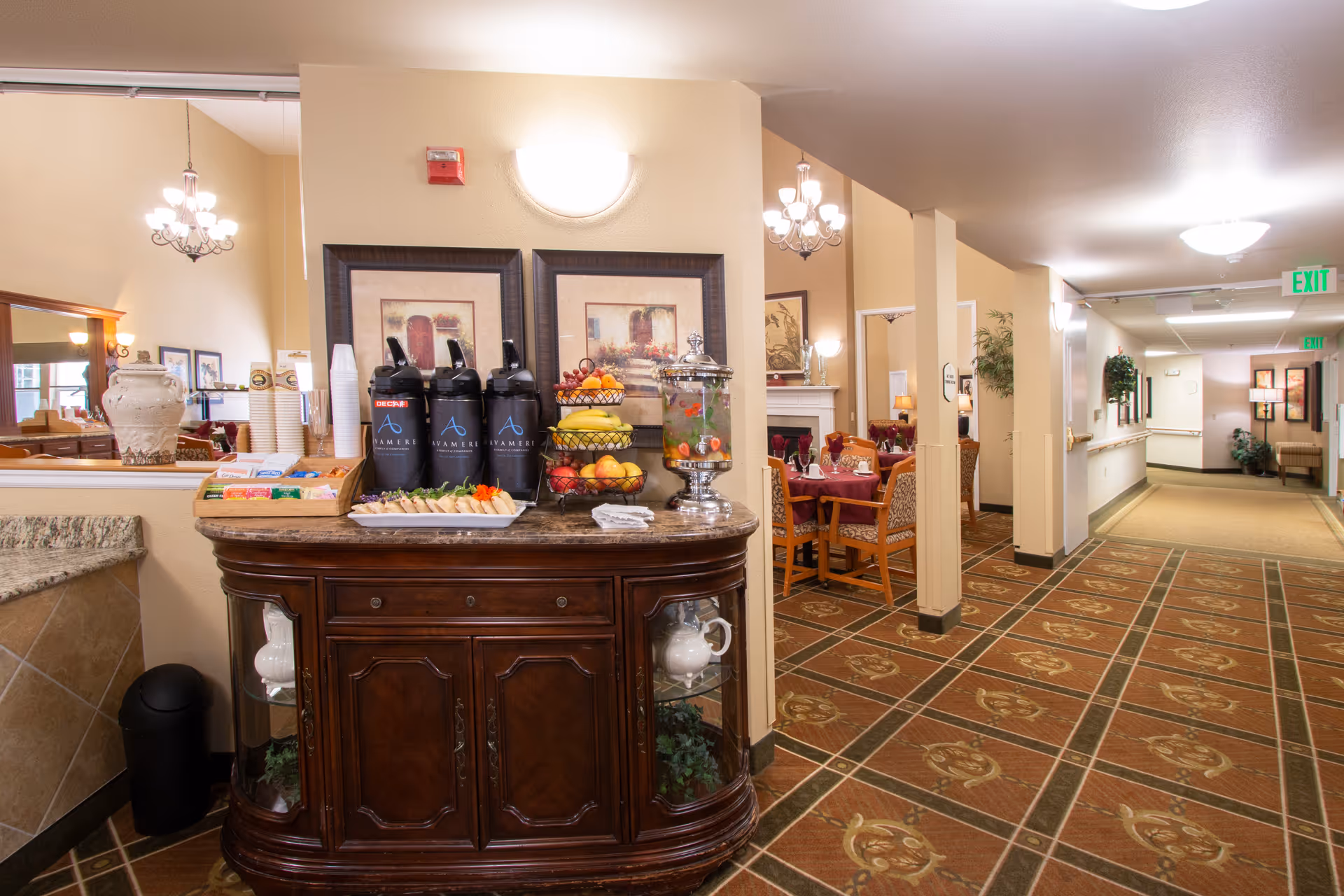 A hallway in a senior living facility with a wooden sideboard holding coffee dispensers, cups, a tray of sandwiches, a tiered fruit basket, and a beverage dispenser with fruit-infused water. The hallway leads to a dining area with tables covered in burgundy tablecloths and chairs. The floor has a patterned carpet, and the walls are decorated with framed artwork and plants.