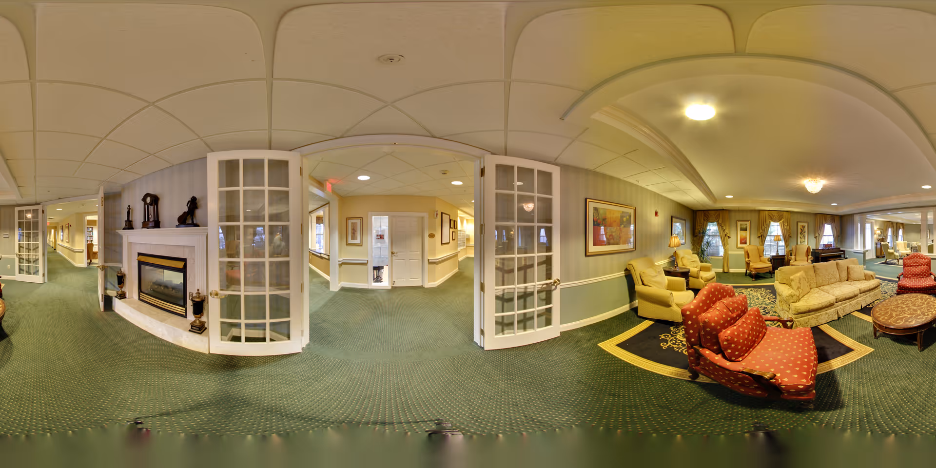 A spacious senior living facility common area with green carpet and beige walls. The room features multiple seating options including red patterned armchairs, beige sofas, and armchairs arranged around coffee tables. There is a fireplace with decorative items on the mantle, framed artwork on the walls, and large windows with curtains allowing natural light. French doors open into a hallway with additional seating and decor.