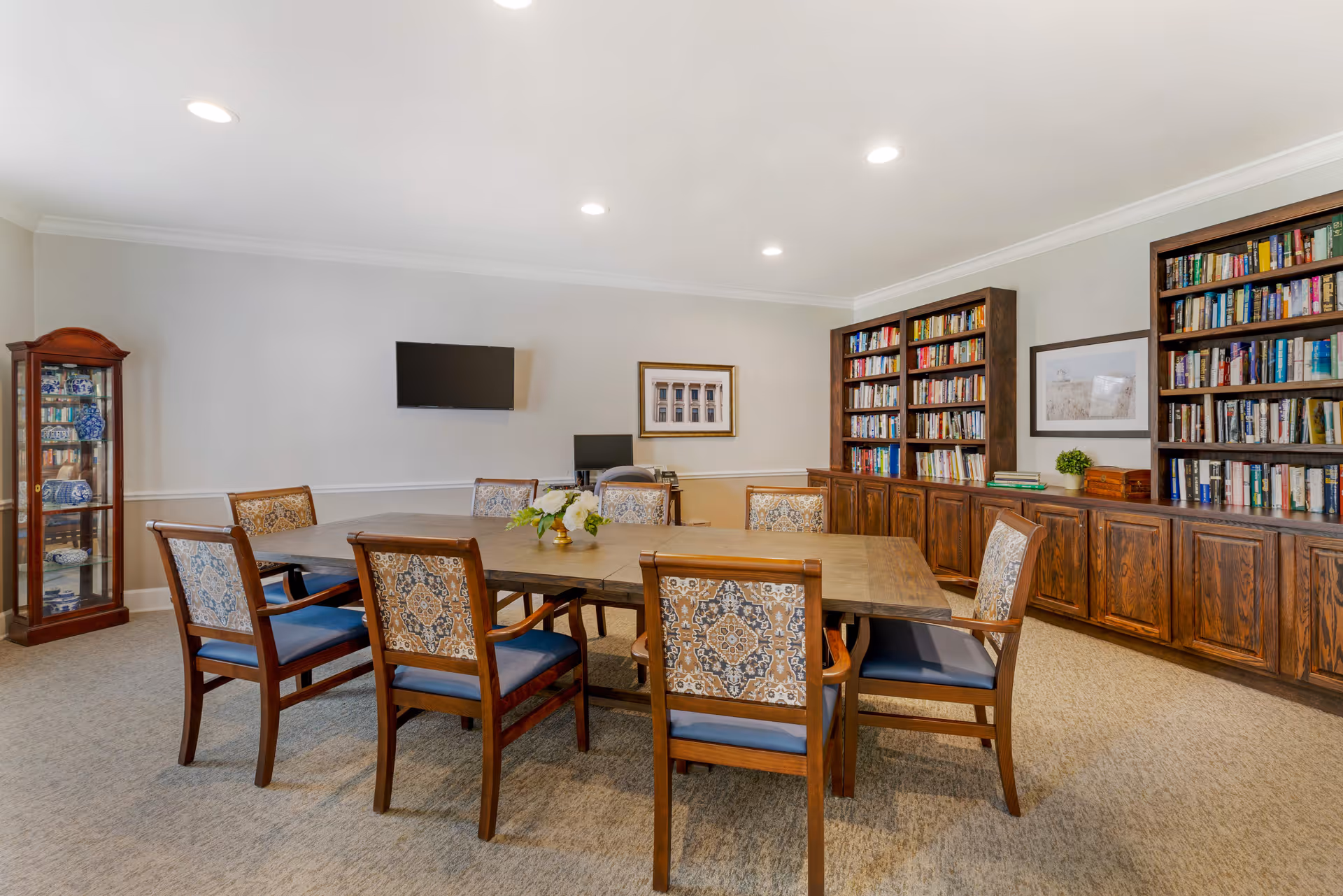 A well-lit room with a large wooden table surrounded by eight upholstered chairs with patterned backs and blue seats. The room features two large wooden bookshelves filled with books, a glass display cabinet with decorative items, a wall-mounted TV, and framed artwork on the walls. The carpeted floor and neutral wall colors create a warm and inviting atmosphere.