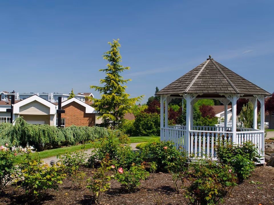 A white wooden gazebo with a shingled roof is situated in a garden area with various green shrubs and blooming flowers. Behind the garden, there are residential-style buildings with brick and siding exteriors under a clear blue sky.