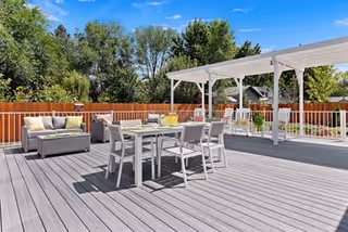Outdoor patio area with gray wooden decking, white dining table and chairs, a seating area with cushioned sofas, and a white pergola. Trees and a wooden fence are visible in the background under a clear blue sky.