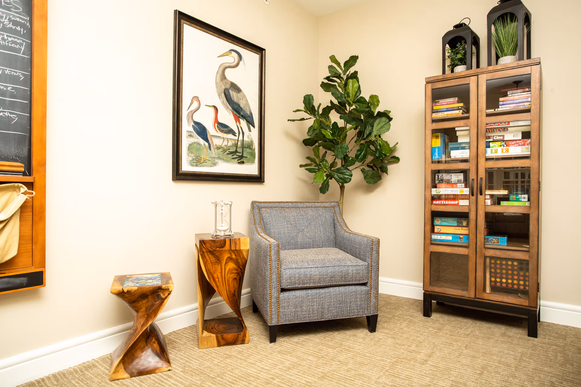 A cozy seating nook with a gray upholstered armchair, two sculpted wooden side tables, a tall cabinet filled with books and games, a potted plant, and framed bird artwork on the wall.