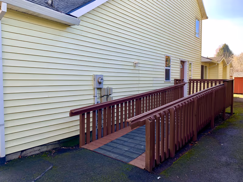 Exterior view of a pale yellow building with a wooden accessibility ramp leading to a side entrance.