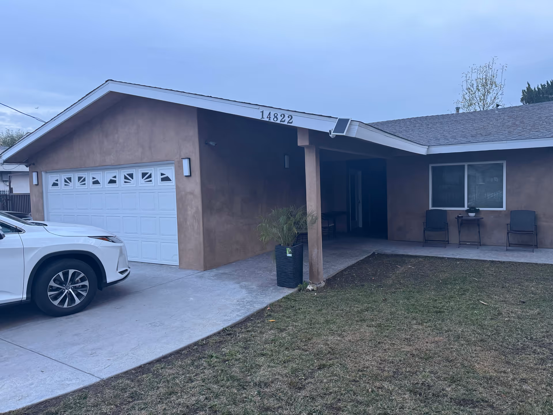 Front exterior view of a single-story building with a brown stucco finish, a white garage door, and a covered porch area with two chairs and a small table. A white vehicle is parked in the driveway, and the house number 14822 is displayed near the roof peak.