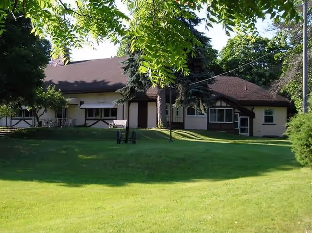 A single-story building with a dark roof and light-colored walls surrounded by green grass and trees, with sunlight filtering through the leaves.