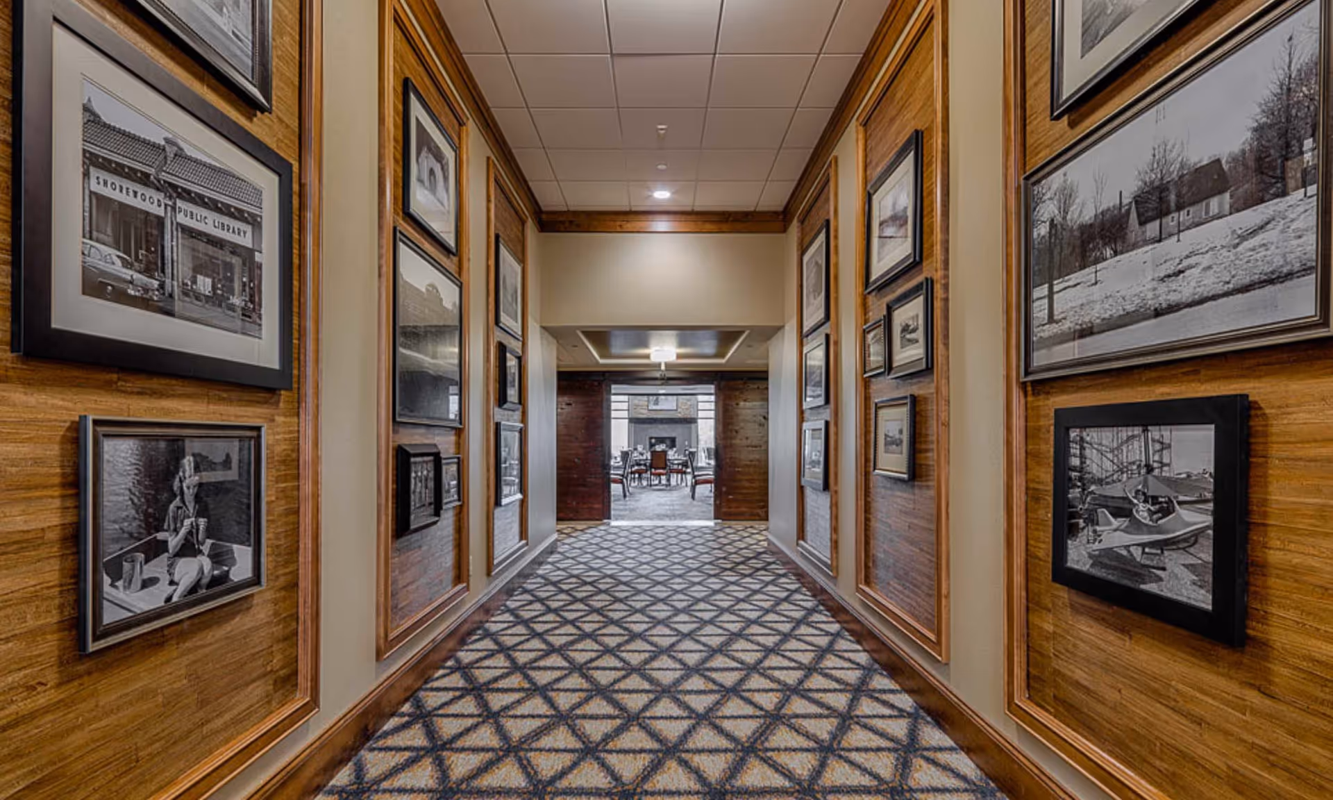 A carpeted hallway with patterned flooring and wooden panel walls adorned with framed black and white photographs. The hallway leads to a room with tables and chairs visible in the background.
