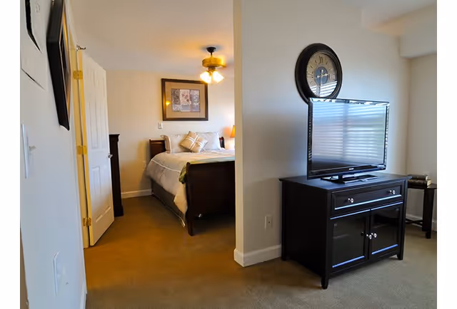 View of a bedroom with a bed, pillows, and a ceiling fan with lights, seen through an open doorway from an adjacent room with a TV on a black cabinet and a large wall clock above it.