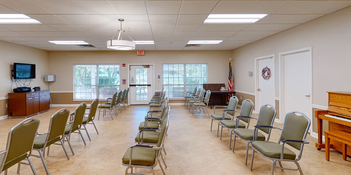 A spacious room with rows of green cushioned chairs arranged facing each other, a wooden piano on the right, a flat-screen TV mounted on the left wall, large windows letting in natural light, and an American flag in the back corner. The room has beige walls and a light-colored floor, with a ceiling light fixture and an exit door at the far end.