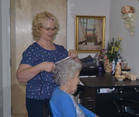 A woman with blonde hair and glasses is styling the gray curly hair of an elderly woman seated in a chair inside a room. The room has a wooden door, a framed painting on the wall, and a table with various decorative items and flowers.