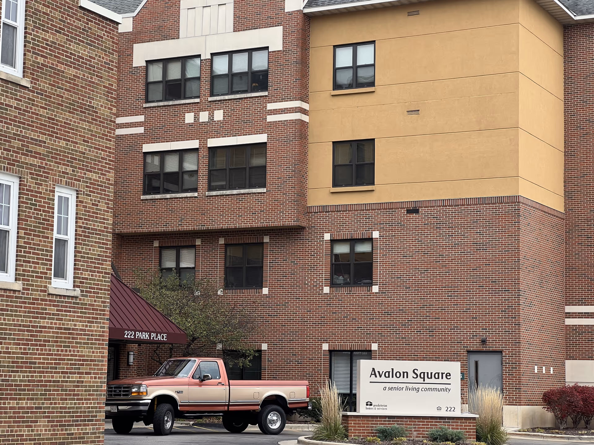 Exterior view of Avalon Square senior living community building with brick and beige facade, a red and beige pickup truck parked in front, and a sign that reads 'Avalon Square a senior living community' near the entrance at 222 Park Place.