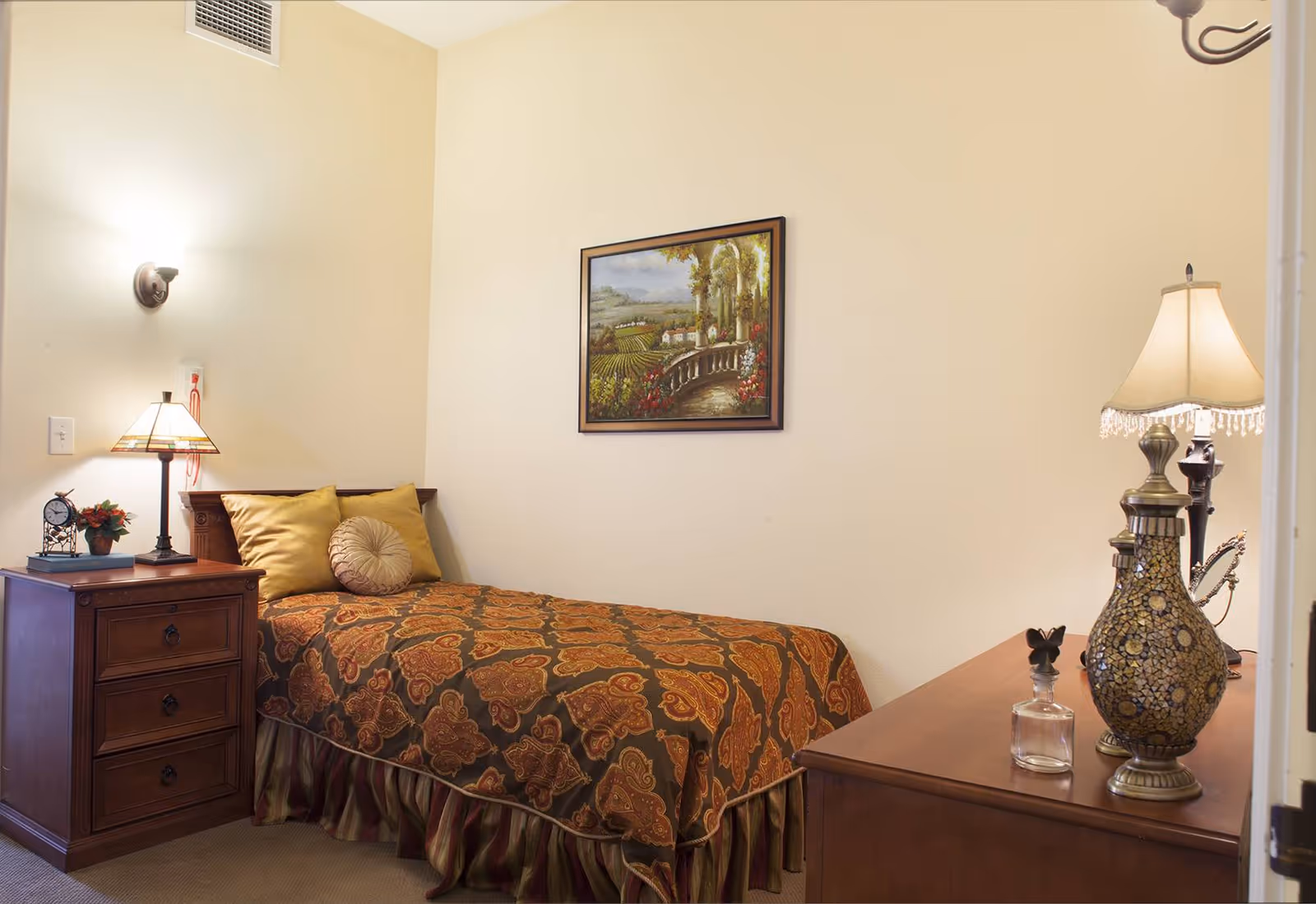 A cozy bedroom with a single bed covered in a patterned brown and orange bedspread, two pillows, a wooden nightstand with a lamp, clock, and small plant, and a wooden dresser with decorative lamps and a small glass bottle. A framed landscape painting hangs on the beige wall above the bed.