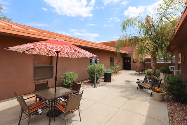 Outdoor courtyard area at Coronado Healthcare Center with a table and four chairs under a red patterned umbrella, benches, potted plants, palm trees, and a clear blue sky.