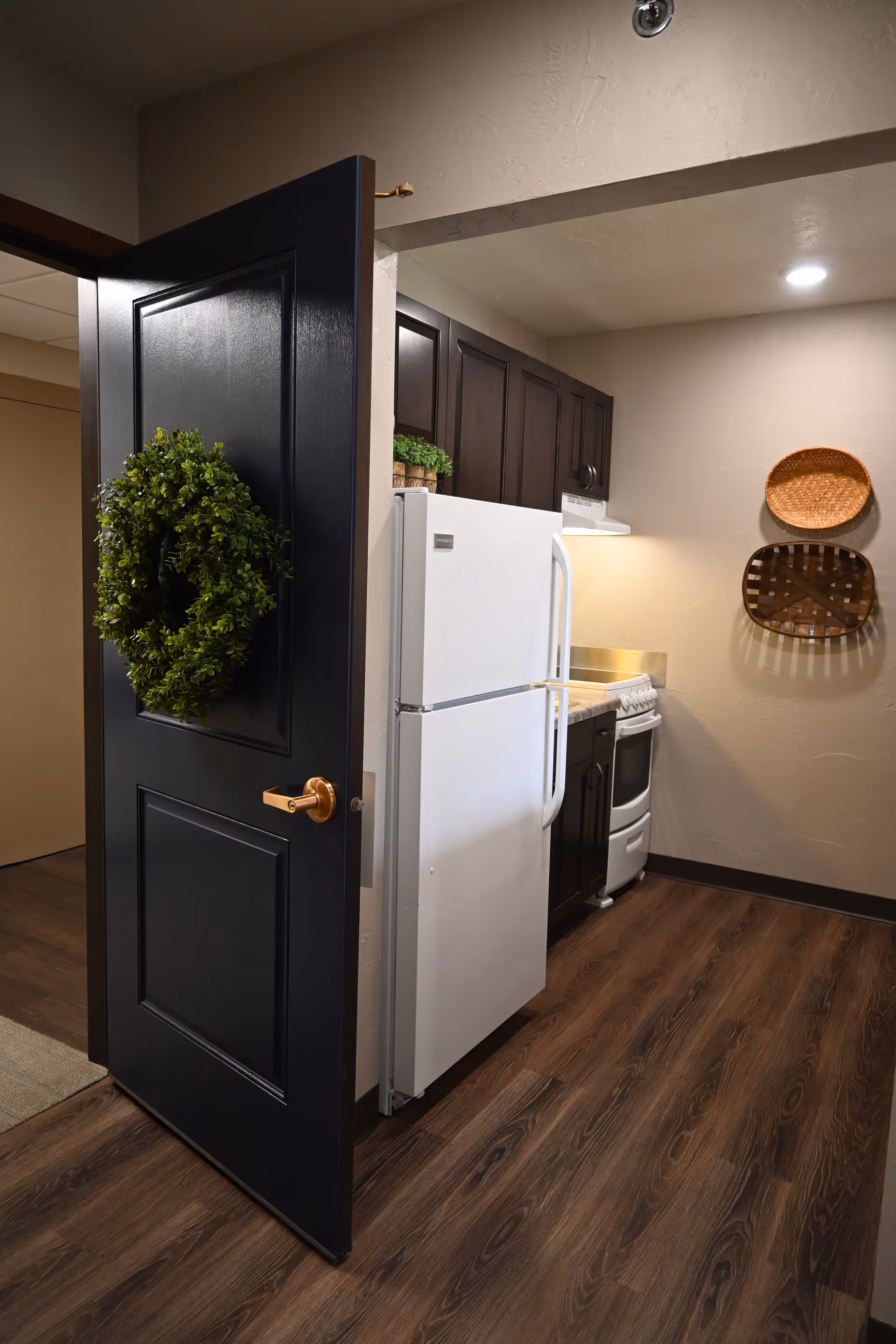 Small kitchen with a white refrigerator and stove, dark cabinets, wood flooring, and a black door hung with a green wreath.