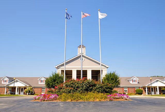 Front entrance of a single-story brick senior living facility with three flagpoles, landscaped flower beds and a clear blue sky.