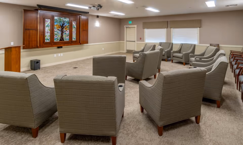 Carpeted common room with rows of upholstered armchairs facing a wood-paneled wall-mounted screen featuring stained-glass artwork.