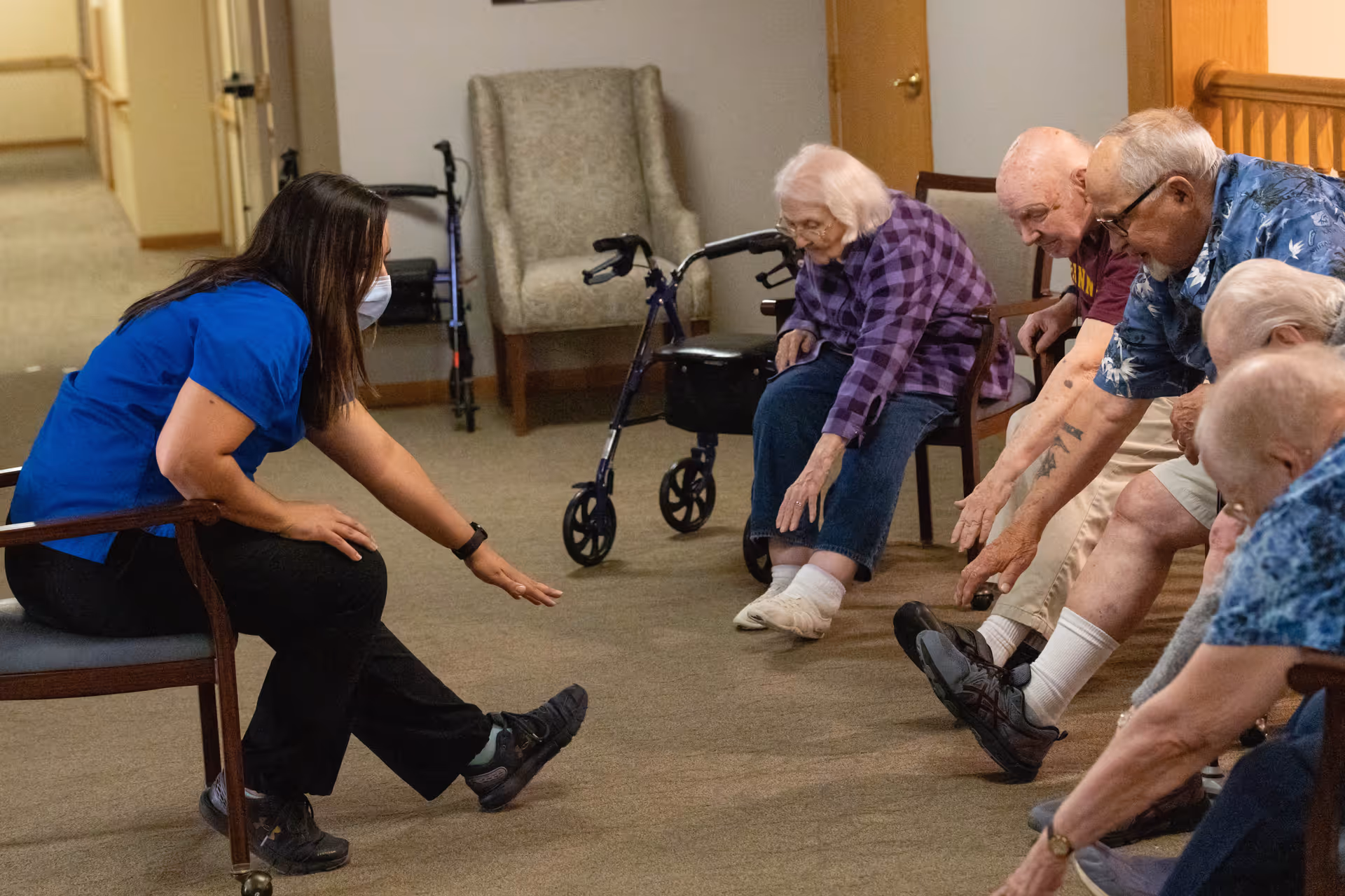 A caregiver wearing a blue shirt and face mask leads a seated group of elderly people in a stretching exercise inside a senior living facility. The elderly participants are seated in chairs and reaching toward their feet. A walker and an armchair are visible in the background.