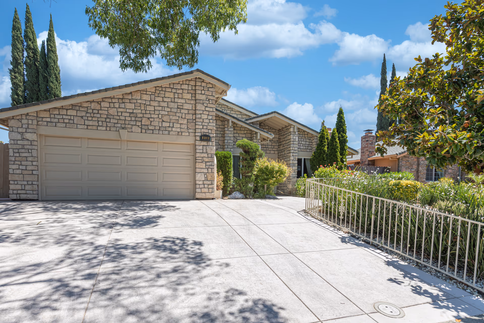Front exterior of a stone-faced single-story home with a two-car garage, driveway, and landscaped yard under a blue sky.