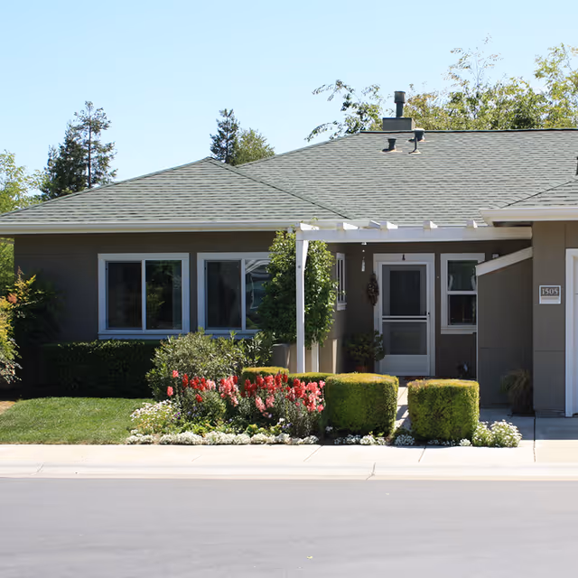 Exterior view of a single-story residential building with a gray roof and beige walls. The entrance features a small covered porch with a white pergola. There are neatly trimmed bushes and colorful flowers in front of the building, along with a well-maintained lawn. Trees are visible in the background under a clear sky.