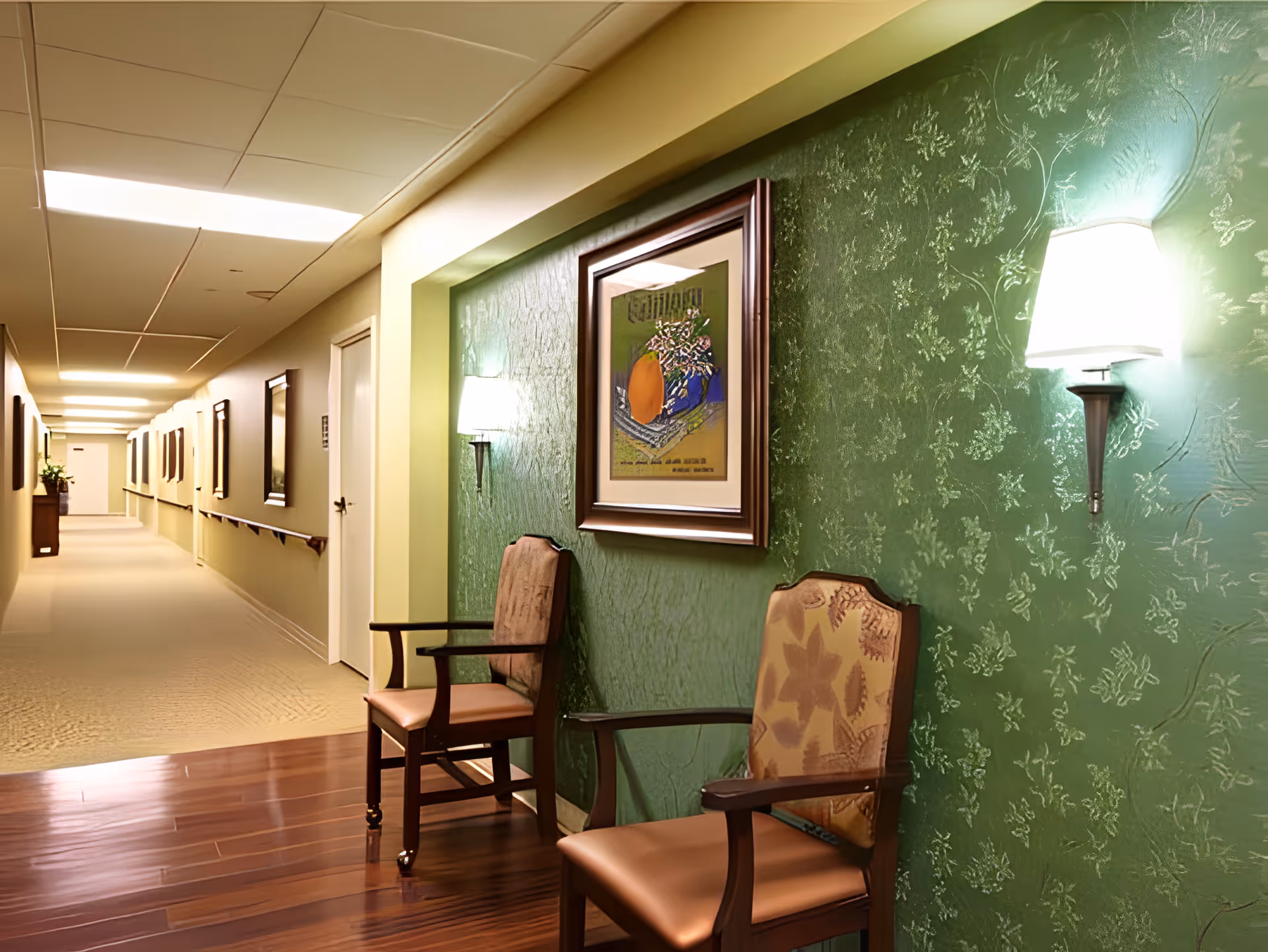 A well-lit interior hallway with two upholstered chairs against a green patterned wall, framed artwork, wall sconces, and a long corridor with handrails.